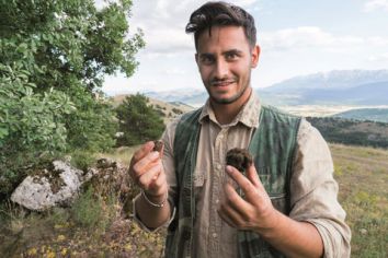 Un giovane uomo sorride mentre tiene un pezzo di terra ricoperto di vegetazione in un ambiente montano.