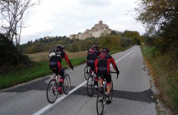 Un gruppo di ciclisti si allena in un paesaggio tranquillo con un antico borgo che si intravede sullo sfondo.