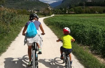 Una madre e un bambino pedalano su un sentiero nella natura, circondati da campi verdi e colline.