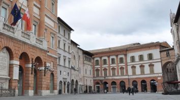 Una scena di una piazza storica in Umbria, caratterizzata da architetture semplici e bandiere che sventolano.