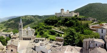 Panorama di un borgo storico con una chiesa e un antico palazzo, circondato da colline verdi.