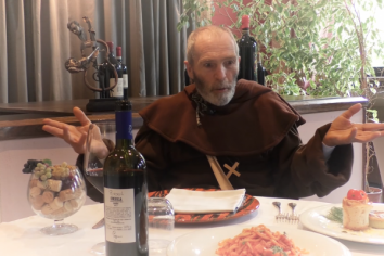 Un uomo vestito da frate discute accanto a un tavolo dove sono serviti vino e cibo.