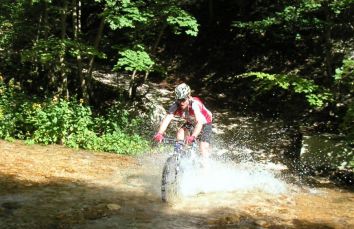 Un ciclista attraversa un torrente in una giornata di sole, spruzzando acqua intorno a sé. La vegetazione verde circostante crea un'atmosfera naturale.