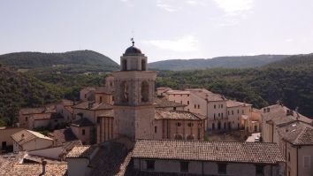 Uno scorcio di un villaggio storico con edifici in mattoni e un campanile che si innalza sopra un paesaggio collinare.