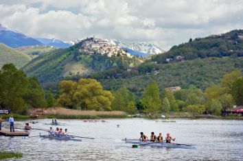 Due equipaggi di canottaggio sul lago, con un borgo storico sullo sfondo e montagne innevate all'orizzonte.