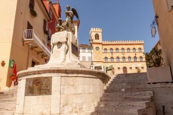 Monumento con angelo in una piazza storica, circondato da palazzi colorati e elementi di architettura antica.