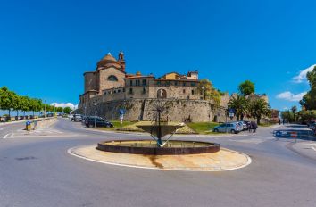 Un castello si erge circondato da un paesaggio verde e ben curato, con una fontana centrale che arricchisce la scena.