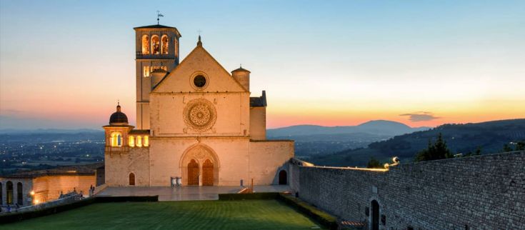 Weekend ad Assisi 4 giorni: city tour con audioguida, ingresso al Bosco di San Francesco - Vacanze Umbria my Love - Scena della Basilica di San Francesco illuminata dal sole al tramonto, situata in un contesto naturale tranquillo.