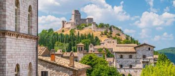 Weekend Umbria 4 giorni. Tour Assisi con audioguida, vista della Rocca Maggiore. Umbria my love - Un paesaggio umbro con una fortezza antica e case in pietra, circondato da vegetazione verde.