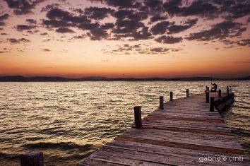 Un molo in legno si estende su un lago tranquillo al tramonto, con due pescatori che attendono di pescare.