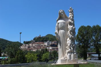 Una statua di una figura religiosa si trova in primo piano, mentre sullo sfondo si vede un borgo umbro situato su una collina verde.