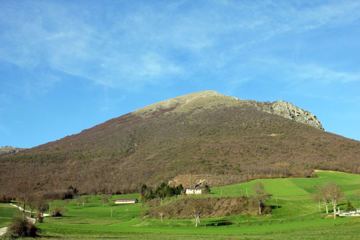 Scena di un monte circondato da ampi campi verdi e un casale in stile rustico.