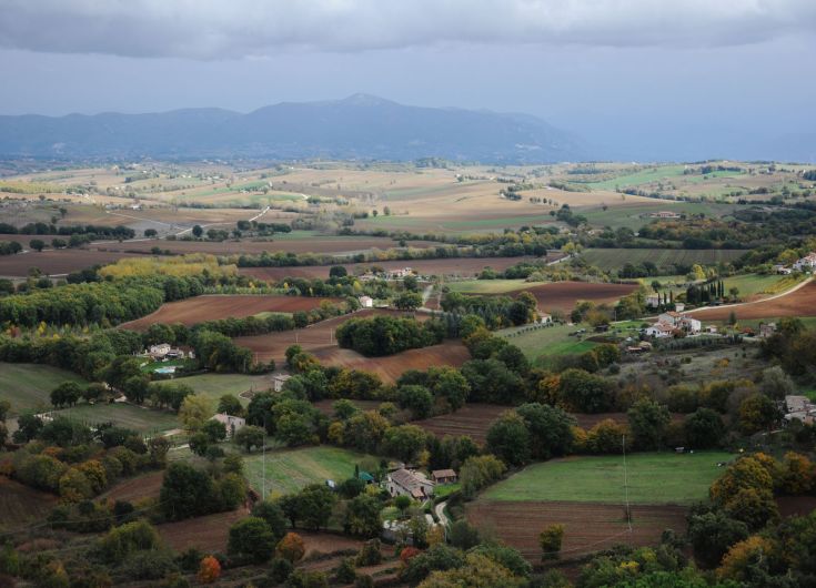 Paysage rural avec des collines, des maisons dispersées et de la végétation verte sous un ciel nuageux.