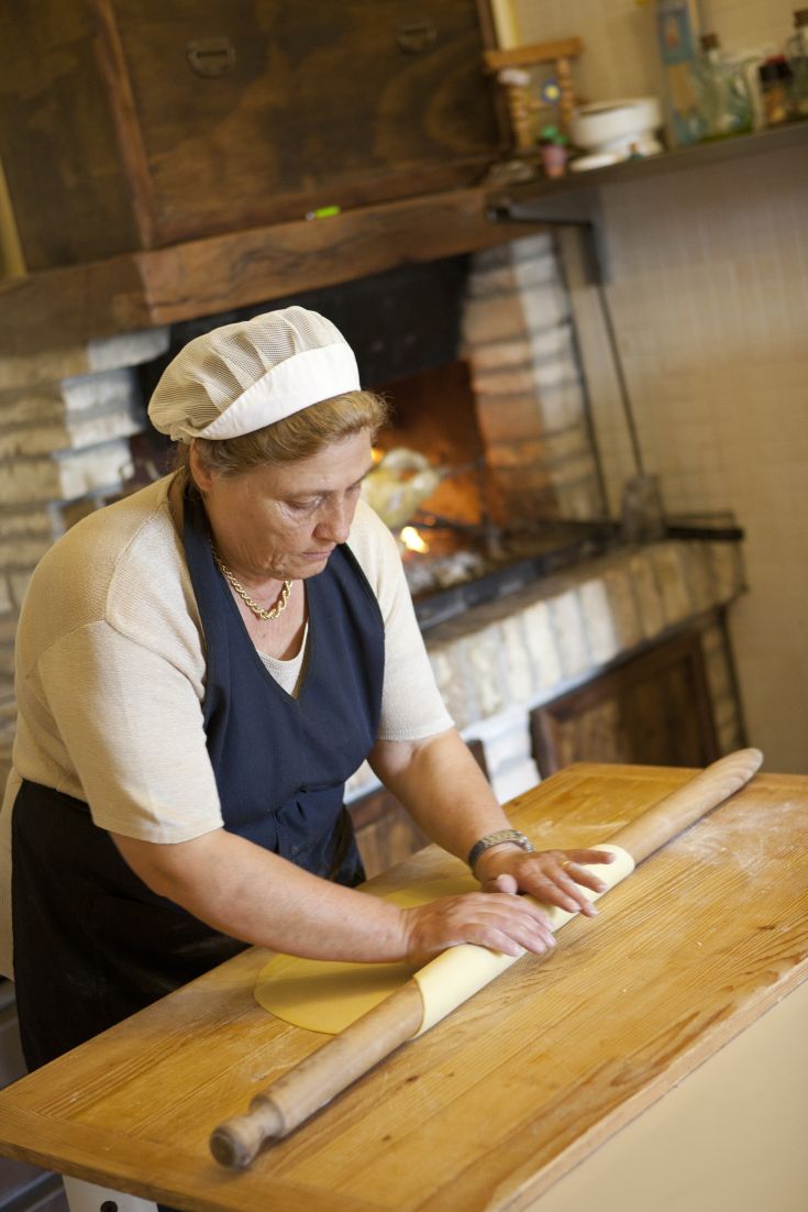 Una donna stende la pasta in una cucina calda, con un camino acceso che crea un'atmosfera accogliente.