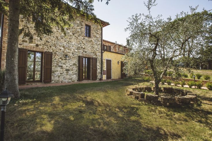Scene of a renovated farmhouse surrounded by greenery, featuring an olive garden and a relaxation area.