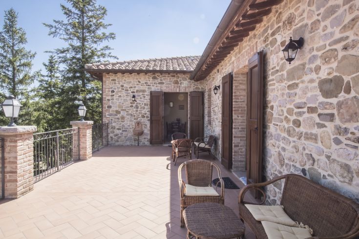 A stone terrace with wicker chairs, surrounded by greenery and trees, on a sunny day.