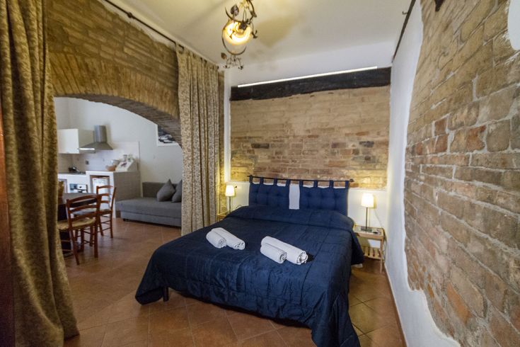 Camera semplice con letto matrimoniale, angolo cottura e particolari in pietra.