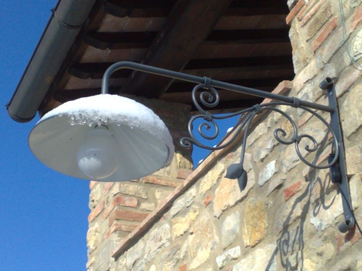 Un lampione semplice ricoperto di neve, montato su una parete in pietra sotto un cielo sereno.