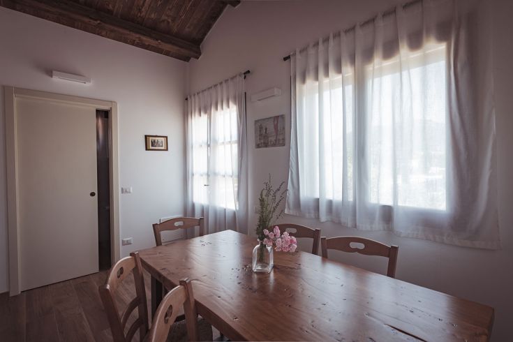 Un ambiente semplice e confortevole con un tavolo in legno e fiori freschi, illuminato da grandi finestre.