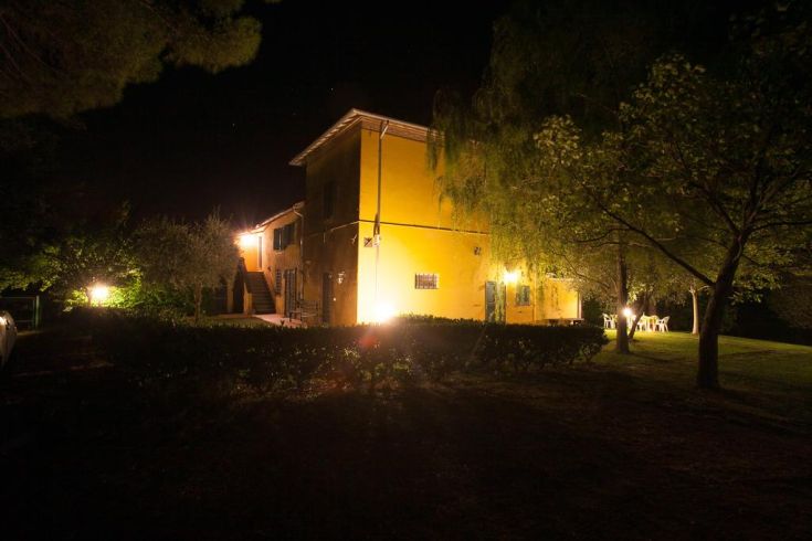 A yellow farmhouse illuminated at night, located in a peaceful area surrounded by greenery.