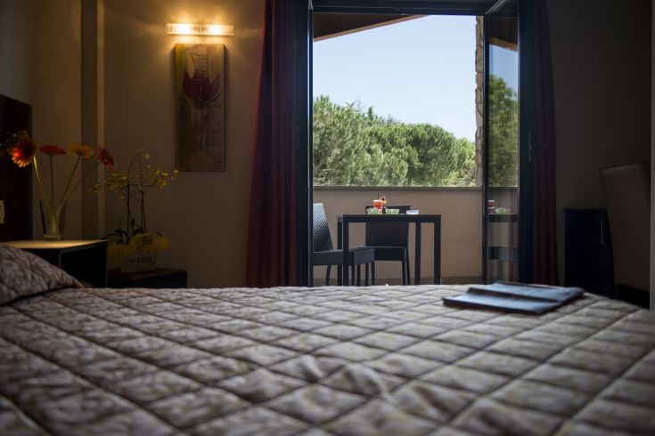 Simple furnished room overlooking a green area, with a small outdoor relaxation corner.