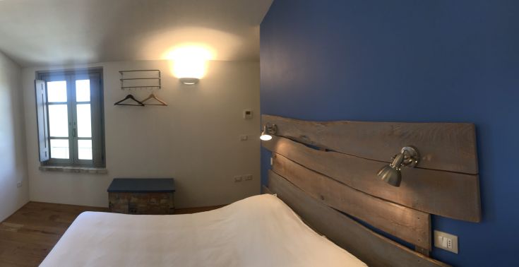 Camera semplice con letto matrimoniale, finestra che illumina l'ambiente e pareti color blu. Spazio accogliente e luminoso.