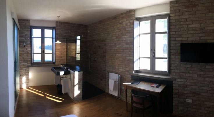 Camera luminosa con pareti in mattoni, arredamento semplice e panorama sulla campagna circostante.