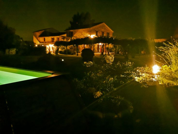 Scène nocturne d'un bâtiment éclairé, entouré d'un jardin et d'une piscine.