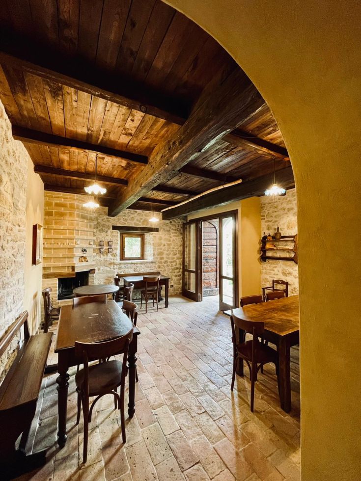 Interno di un palazzo in pietra con tavoli in legno e travi a vista, atmosfera accogliente e semplice.