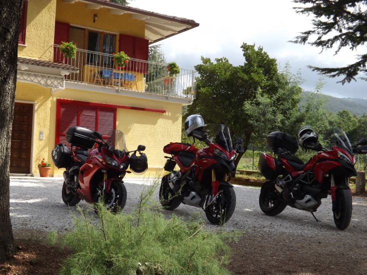 Drei Motorräder vor einem gelben Gebäude, umgeben von Grünflächen.