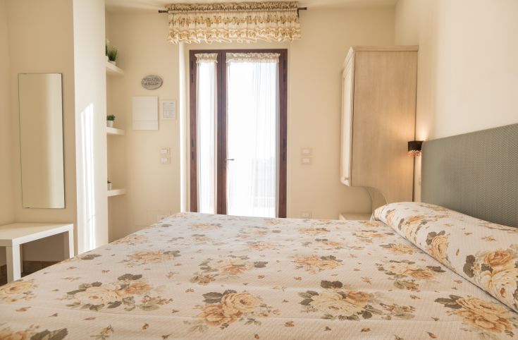 Camera semplice con un letto floreale, illuminata dalla luce naturale e con apertura su una zona verde.