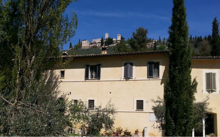 Scene of a countryside house in Umbria, surrounded by greenery with a building in the distance.