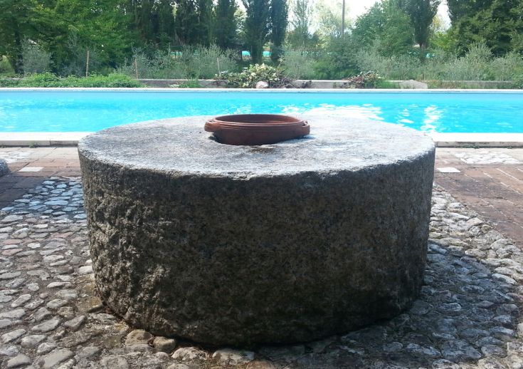 A sturdy circular stone with a terracotta vase, surrounded by greenery near a lake.