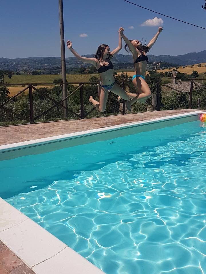 Due ragazze saltano in piscina, immerse in un contesto di natura verde e cieli sereni.