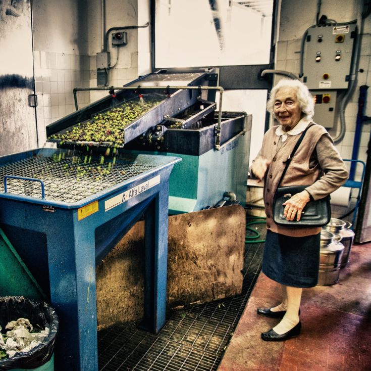 Eine ältere Frau lächelt neben einer Olive-Verarbeitungsmaschine.