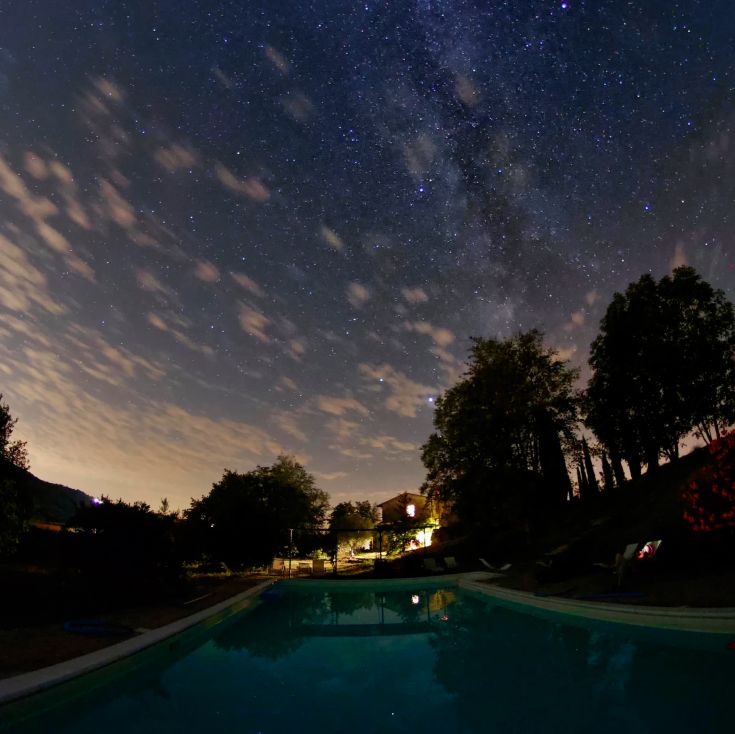 Eine ruhige Nacht über einem Bauernhaus mit einem Pool, der den sternenklaren Himmel und einige leichte Wolken spiegelt.