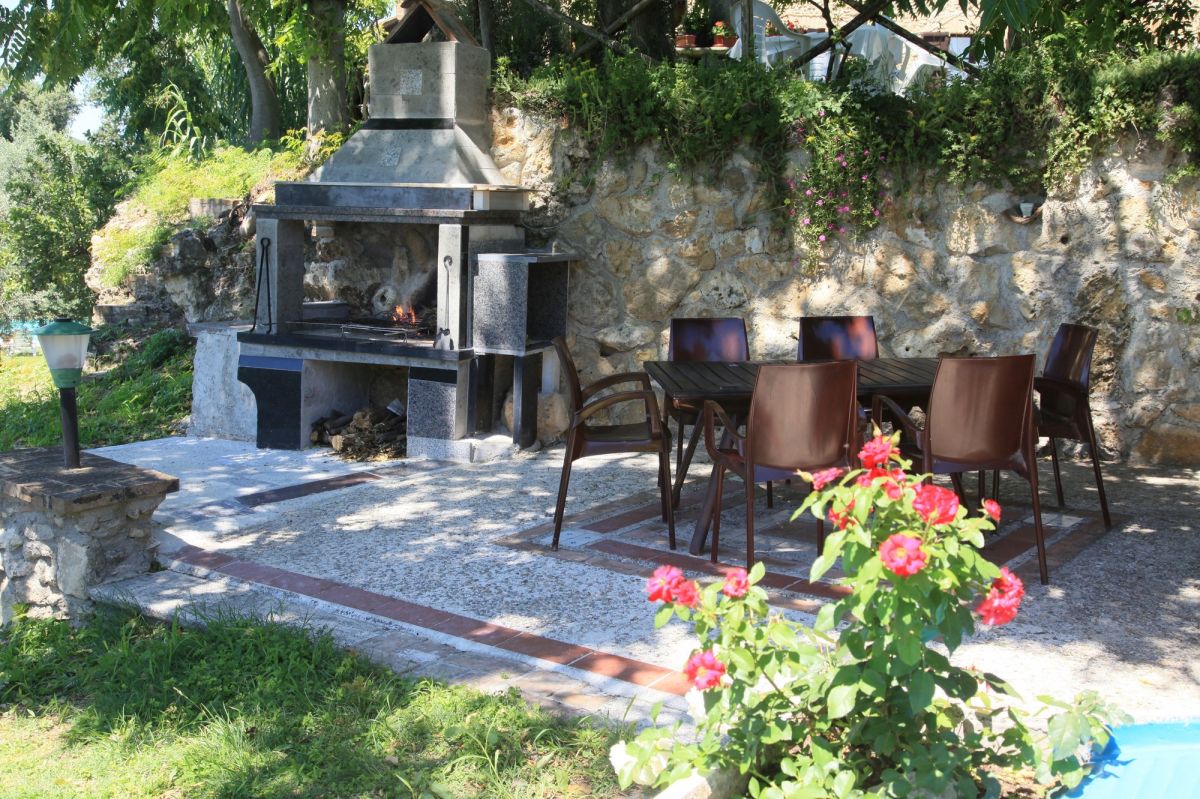 An outdoor green area with barbecue and tables, surrounded by flowering plants.
