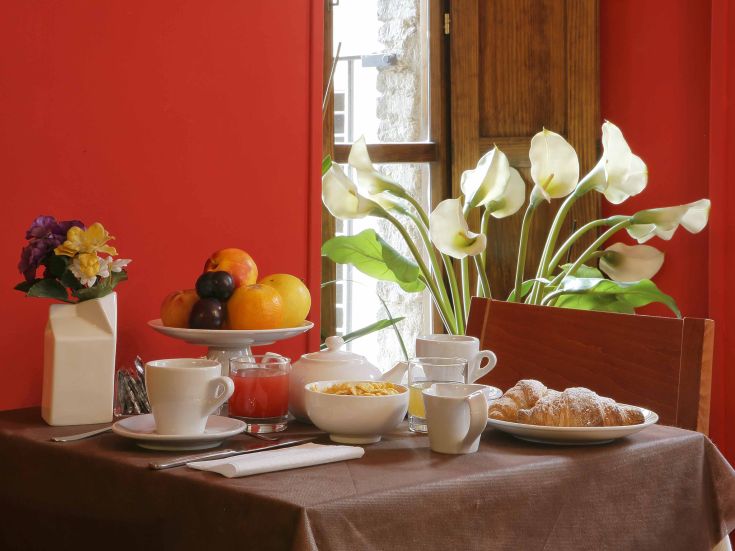 Colazione semplice con frutta, cornetti e bevande, servita in una sala accogliente dai toni rossi.