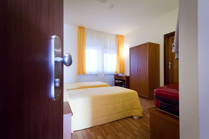 Una camera semplice con letti singoli, armadio e finestre con tende gialle.