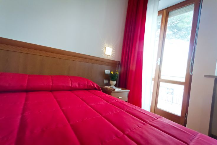 Camera semplice con letti rossi e panoramica su un balcone ben illuminato.