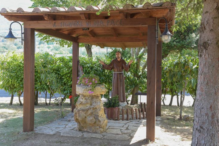 La statua di un santo si trova sotto un ampio portico, circondata da piante verdi che creano un ambiente sereno.