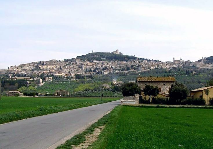 Panorama di Assisi, circondata dalla vegetazione con un cielo blu e sole splendente.