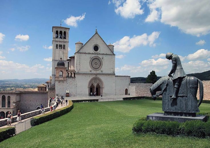 Scena panoramica di Assisi, visibile la basilica e una scultura equestre, circondate da vegetazione.