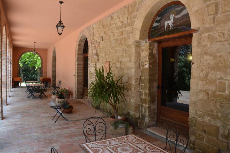 Ein einfacher Portikus einer Villa mit Außentischen, Pflanzen und einer umliegenden grünen Landschaft.