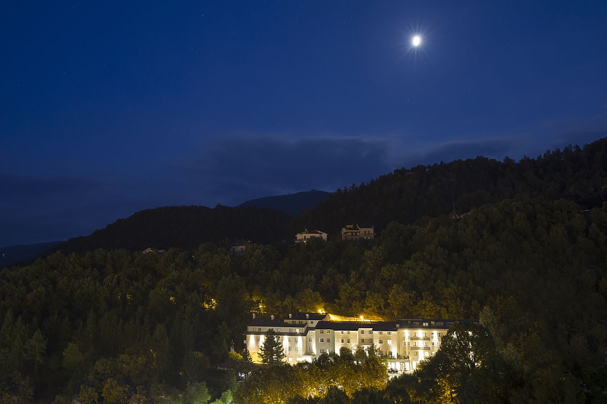 Scena notturna di un hotel circondato da alberi, con la luna illuminante e montagne visibili in lontananza.