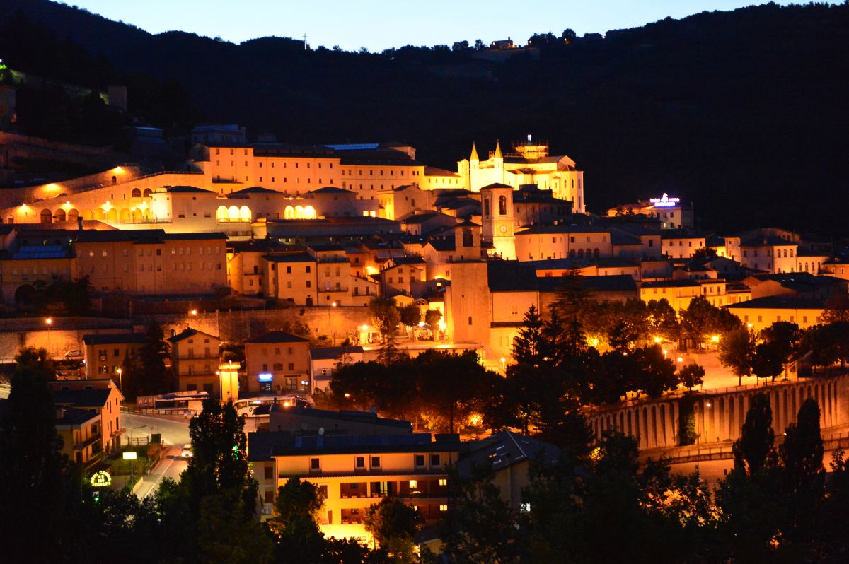 Night scene of a village, featuring illuminated historic buildings and mountains in the background.