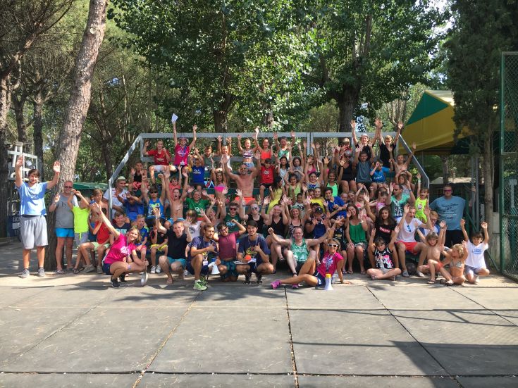 A happy group of children and teenagers smiles and waves, surrounded by green space under the sun.