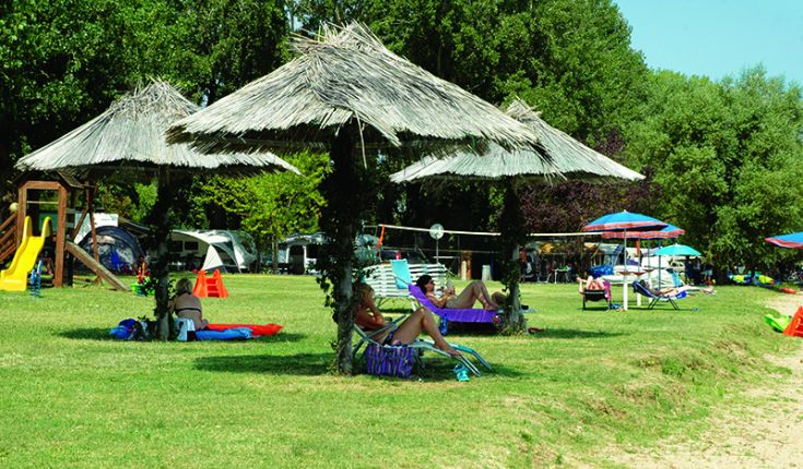 Une scène de camping tranquille au bord du lac, avec des tentes et des parasols entourés d'arbres et de nature.