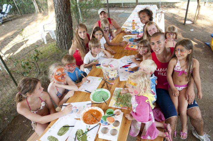 Un gruppo di bambini e adulti si dedica a attività artistiche con colori e pennelli in un ambiente di campeggio.