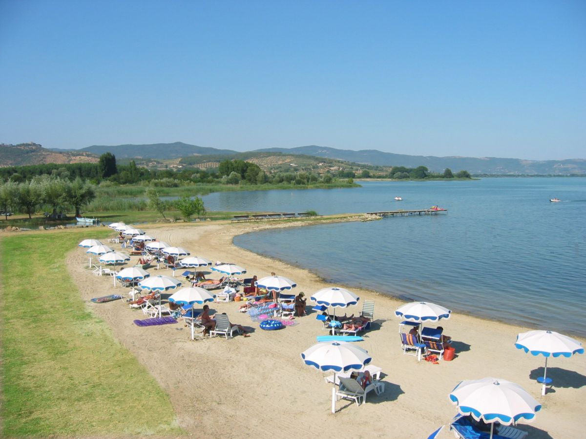 Spiaggia con sdraio e ombrelloni di fronte al Lago Trasimeno, circondata da colline tranquille.